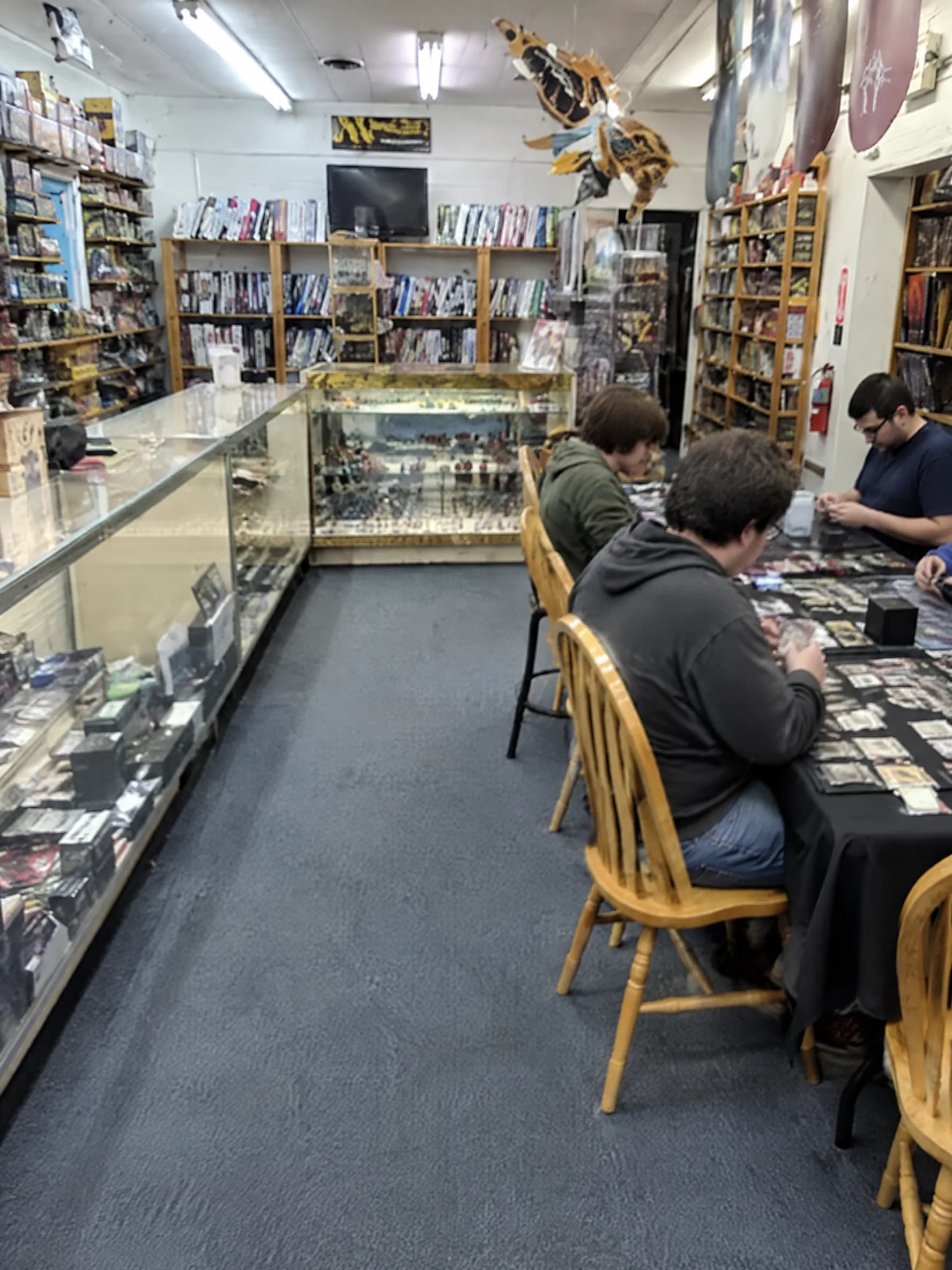 The main aisle at Flights 2: glass card cases on one side, shelves of board games on the other, players at the tables in the back.